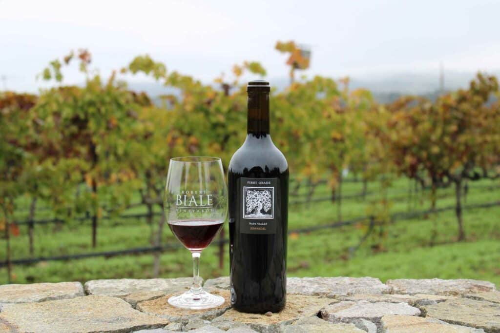 bottle of Biale First Grade Zinfandel beside a filled wine glass, sitting on a rock wall with vines behind