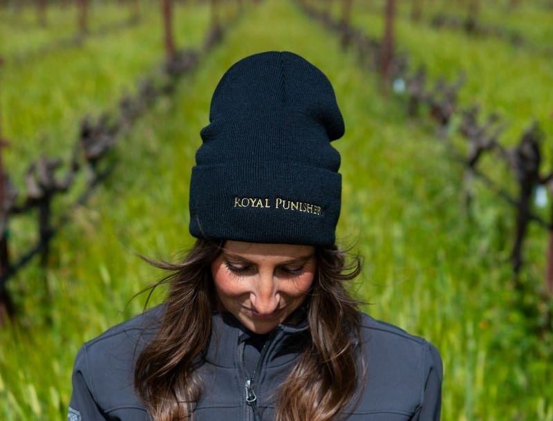 woman looking down while wearing a Royal Punishers beanie with vineyard row behind her